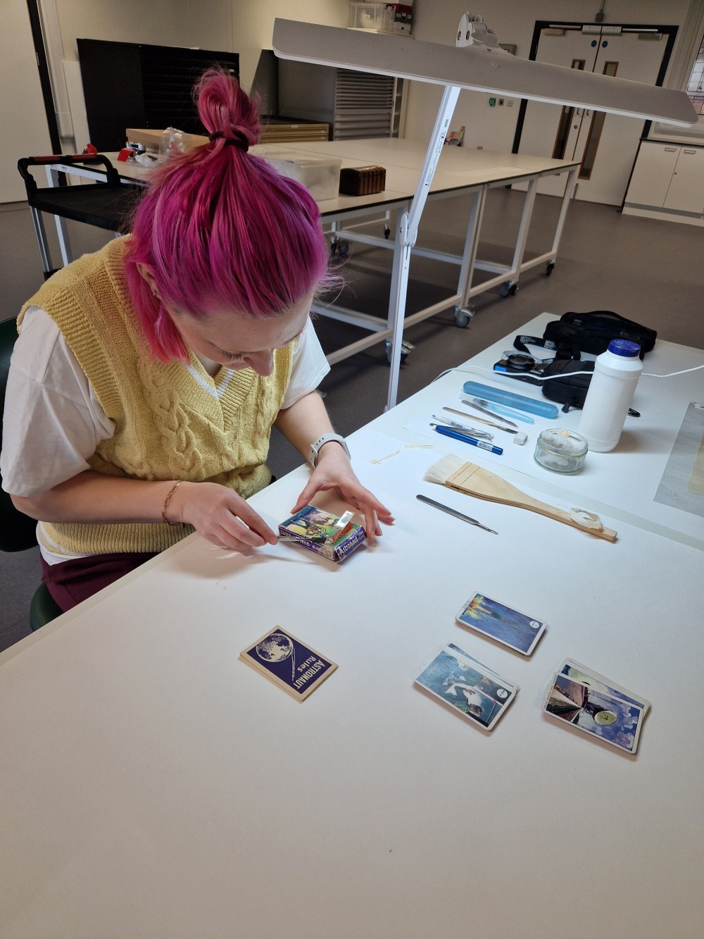 Jessica sits at a white workbench, working on the card game. She has pink hair and wears a yellow knit vest and white tshirt.
