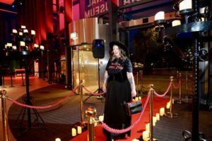 Woman in a gothic outfit of long black skirt, black waistcoat, hat and t'shirt reading 'directed by Terence Fisher' walks up the spooky red carpet into the museum