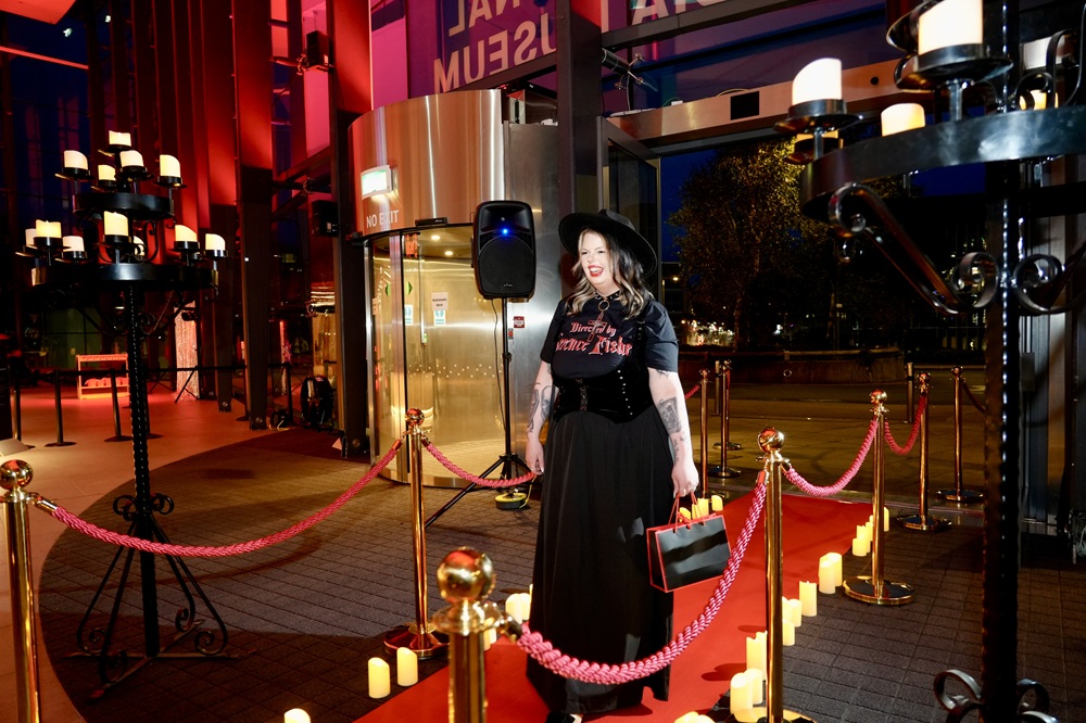 Woman in a gothic outfit of long black skirt, black waistcoat, hat and t'shirt reading 'directed by Terence Fisher' walks up the spooky red carpet into the museum
