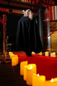 Woman in a black cape walks up a red carpet lined with candles, making a shocked expression