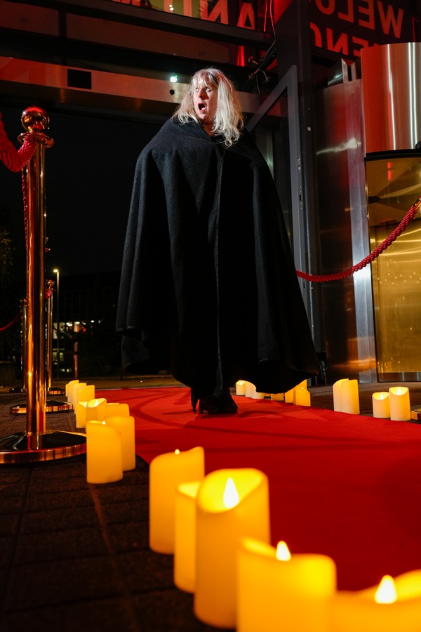 Woman in a black cape walks up a red carpet lined with candles, making a shocked expression