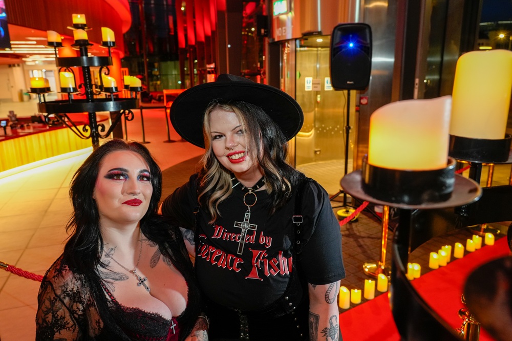 Two women in goth clothing smile at the camera in the museum foyer, which is lit by candles on the floor and in candelabras.