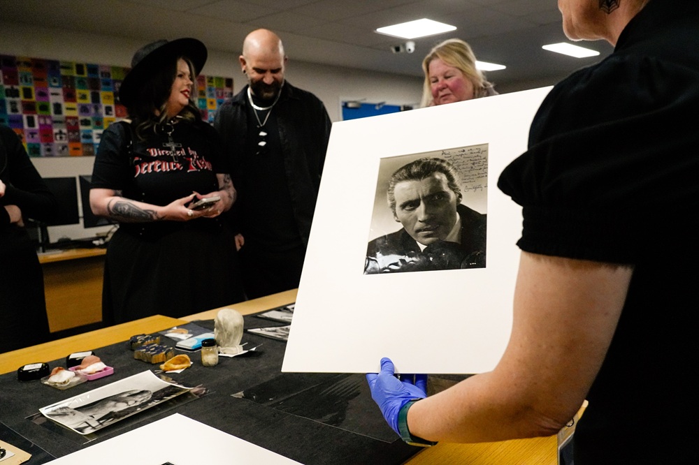Guests look at a display of Hammer-related objects in Insight. I the foreground, a colleague wearing blue nitrile gloves holds up a photograph of Christopher Lee