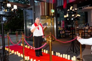 A woman wearing black trousers, white blouse and bright red scarf has her photo taken on the candlelit red carpet