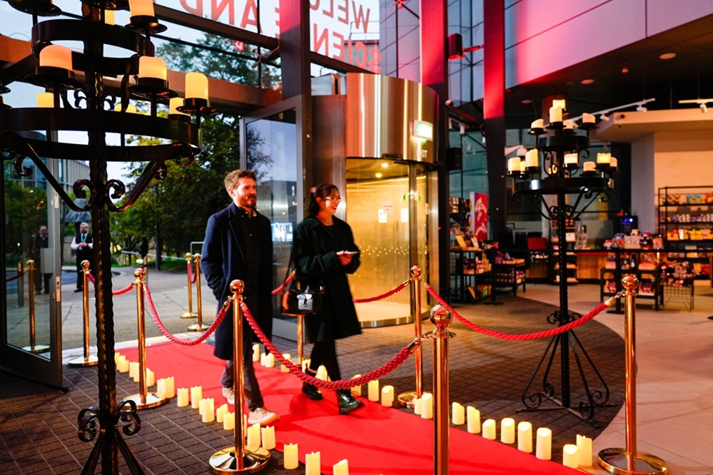 A man and woman in long dark coats walk smiling up the candlelit red carpet into the museum