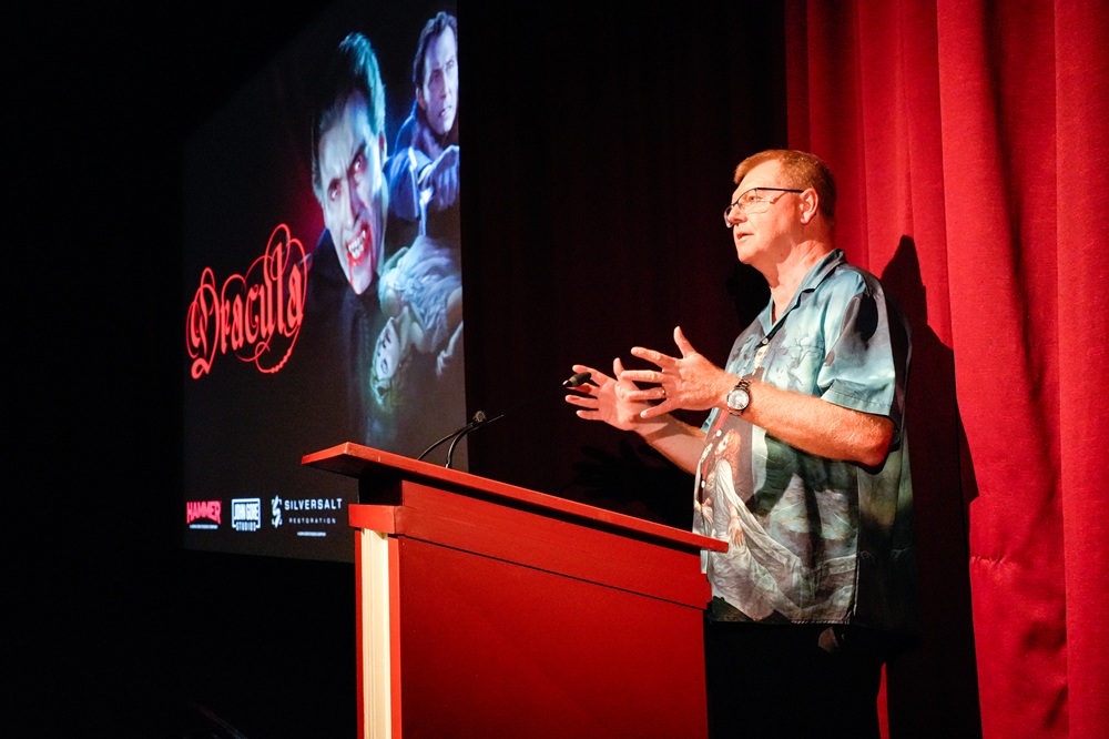 A man wearing a shirt with a Dracula print stands at the lectern in Pictureville, with the red velvet curtain and Dracula poster on screen behind him